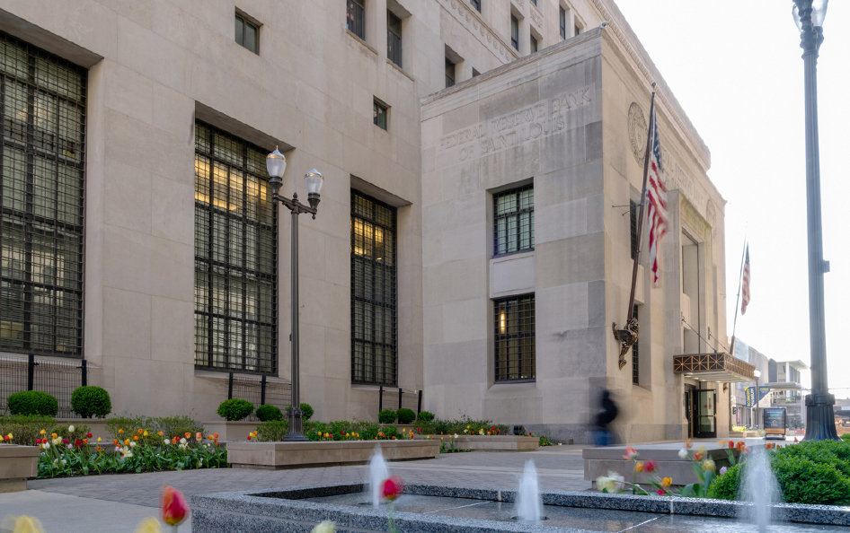 A fountain, paved plaza and flowers are in front of a stone building with U.S. flags.