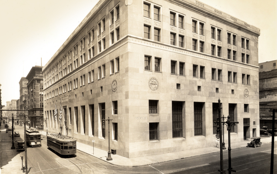 Streetcars pass a white stone building at a downtown intersection in a black and white photo.