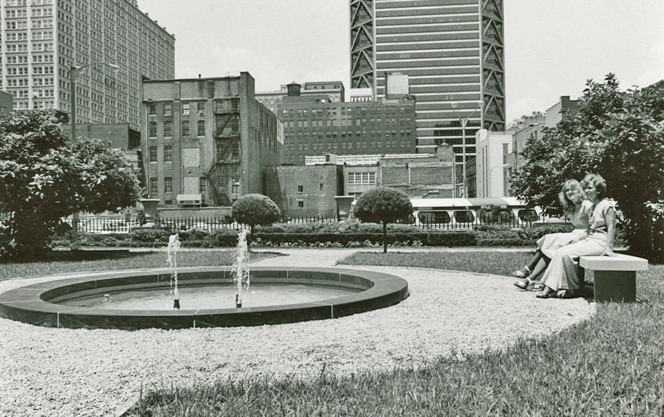 Two women in 1970s-era clothes sit on a bench by a fountain in front of a downtown skyline in a black and white photo.