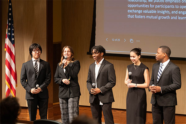 Five interns present to an auditorium filled with people in front of a large screen.