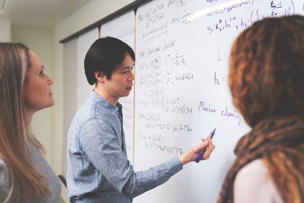 Man writes on a whiteboard as two colleagues observe.