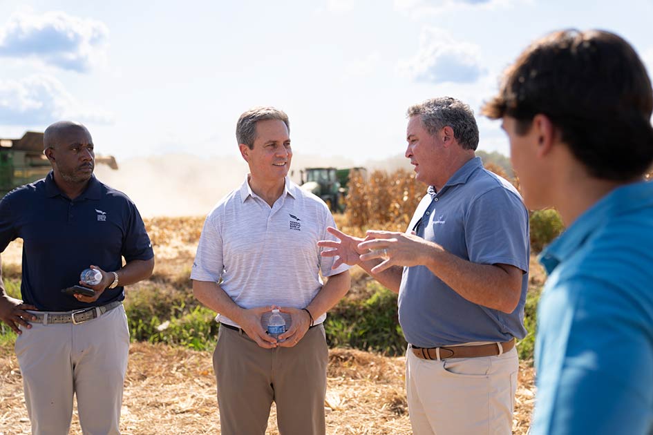 A man speaks to St. Louis Fed President Alberto Musalem and two others while corn is harvested in the background.